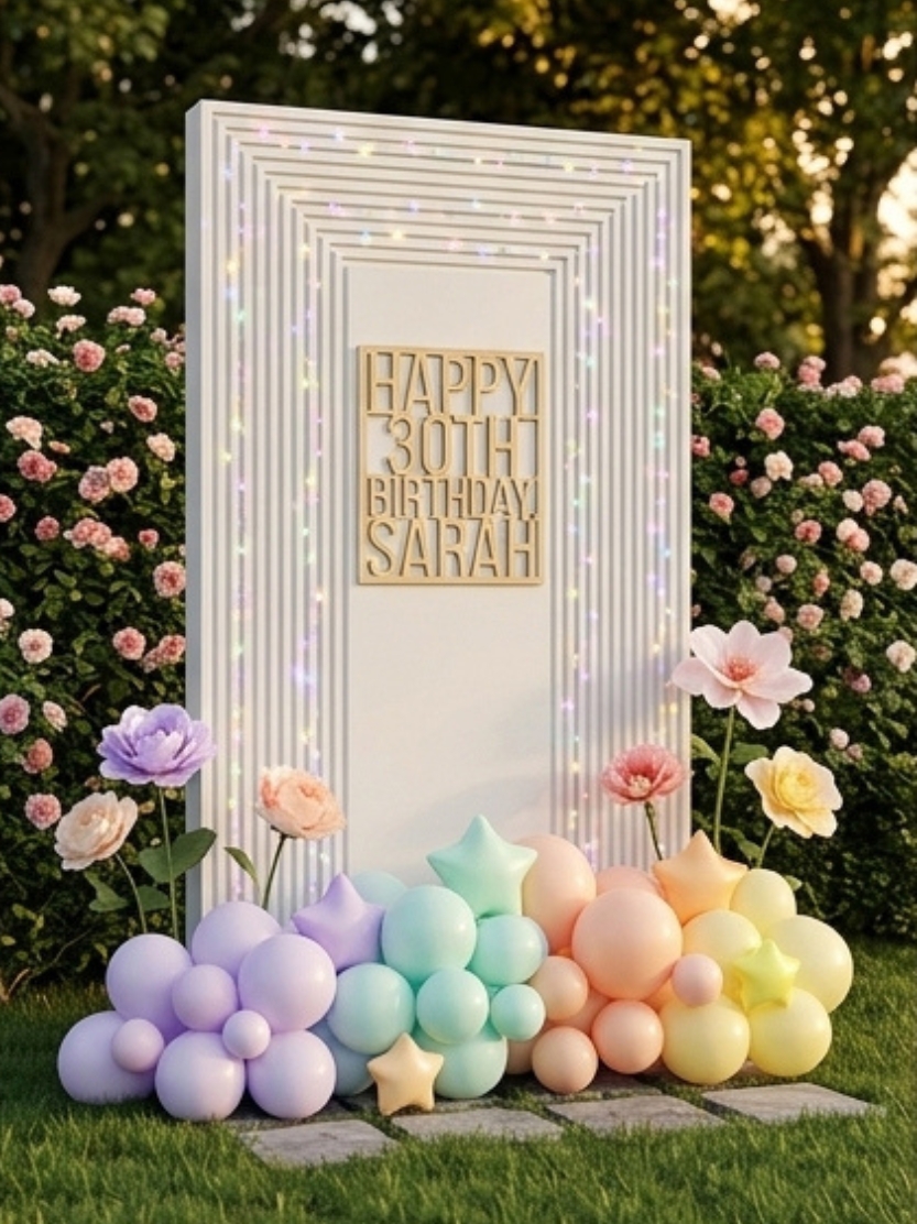 White ribbed moulding frame backdrop panel with fairy lights, a custom wooden Happy 30th Birthday sign, pastel balloon garland, and giant flowers set in an outdoor garden.